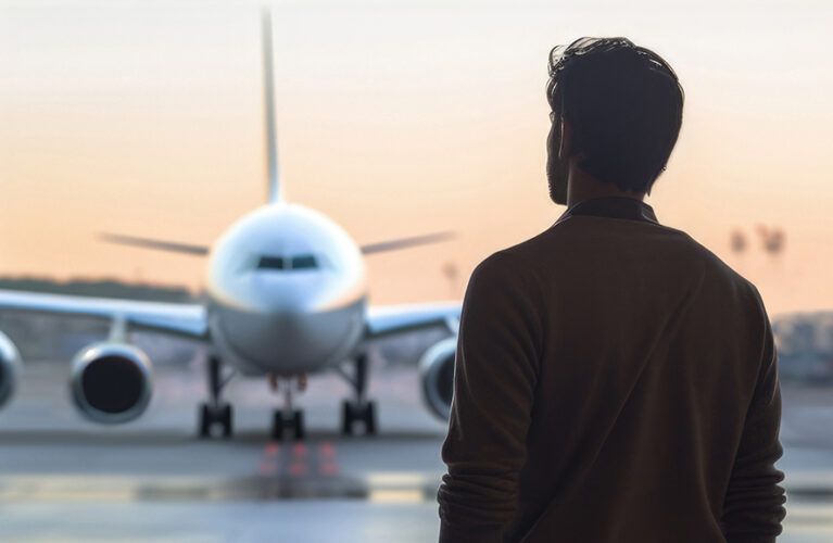 Man standing in an airport terminal looking at a plane ready for takeoff at sunset.